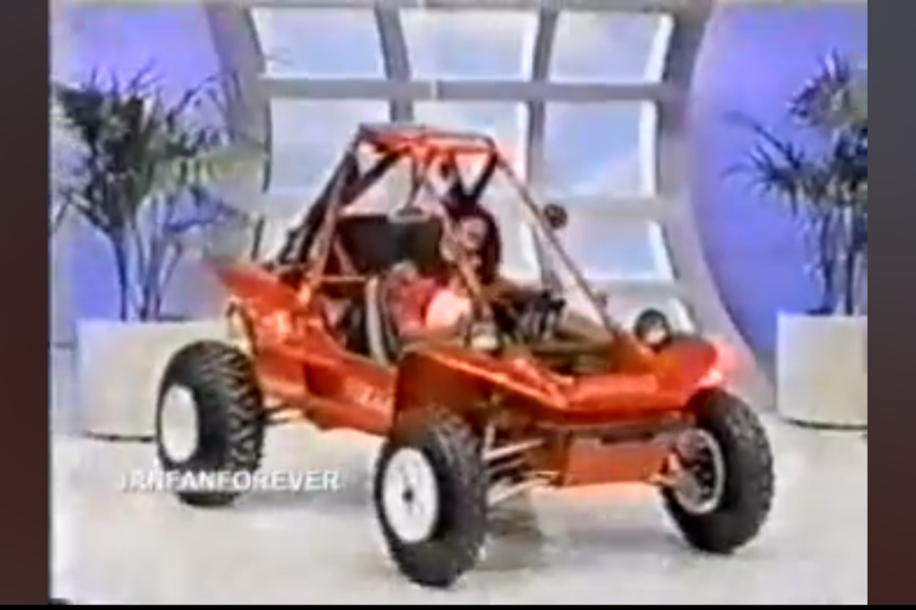 A red dune buggy displayed indoors with a blue and white background, featuring potted plants beside it.