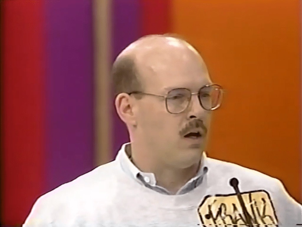 A Price is Right contestant named Frank with glasses and a mustache, wearing a light-colored sweatshirt, stands at a microphone with a surprised expression against a colorful background.