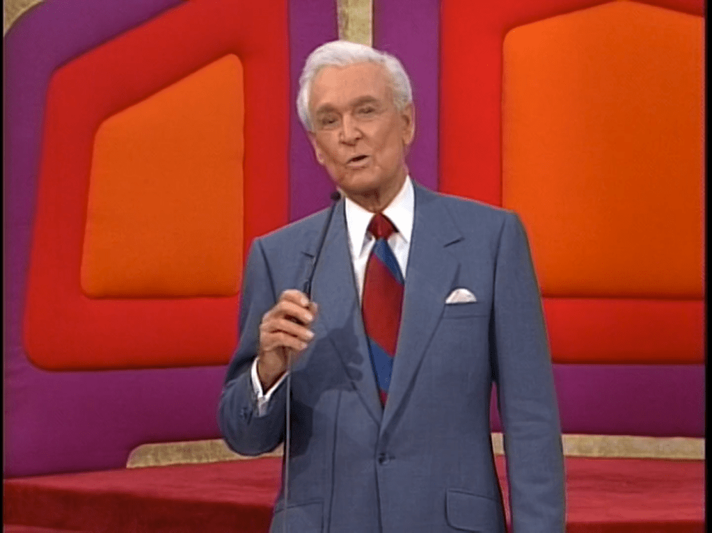 Bob Barker in a gray suit and red, blue, and white tie stands in front of a colorful backdrop, speaking into a microphone.