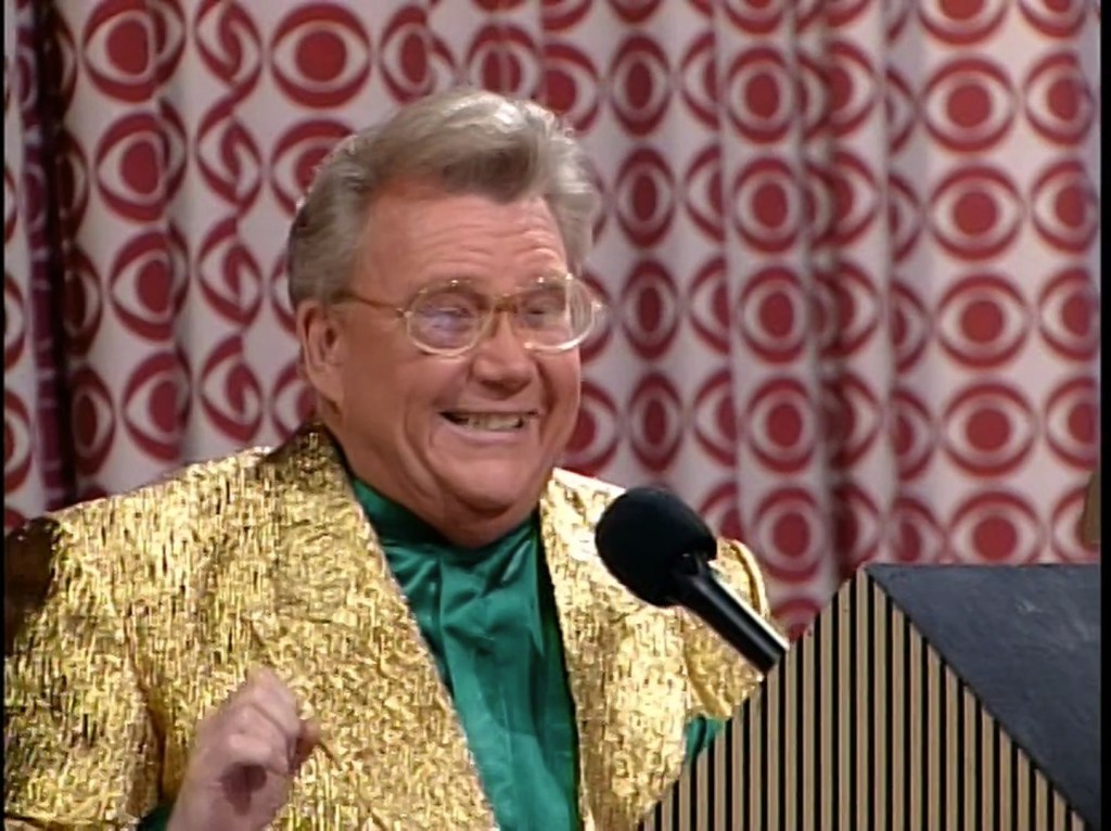 Rod Roddy, a man with light hair and glasses smiles enthusiastically while speaking into a microphone. He is wearing a shiny gold jacket over a green shirt, with a patterned background featuring the CBS eye.