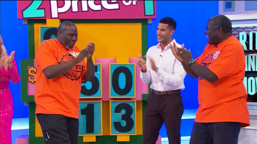 Two contestants in orange shirts celebrating with a host on a game show set, with a colorful backdrop displaying numbers.