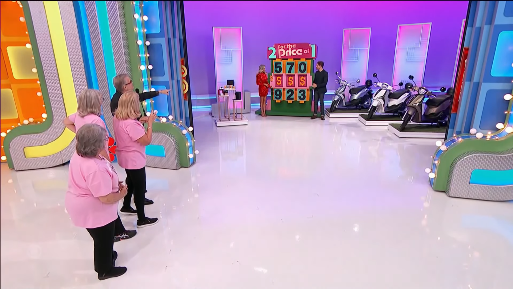 A game show scene with a host pointing, contestants in pink shirts, and a display board featuring the numbers 570 and 923, alongside several scooters.