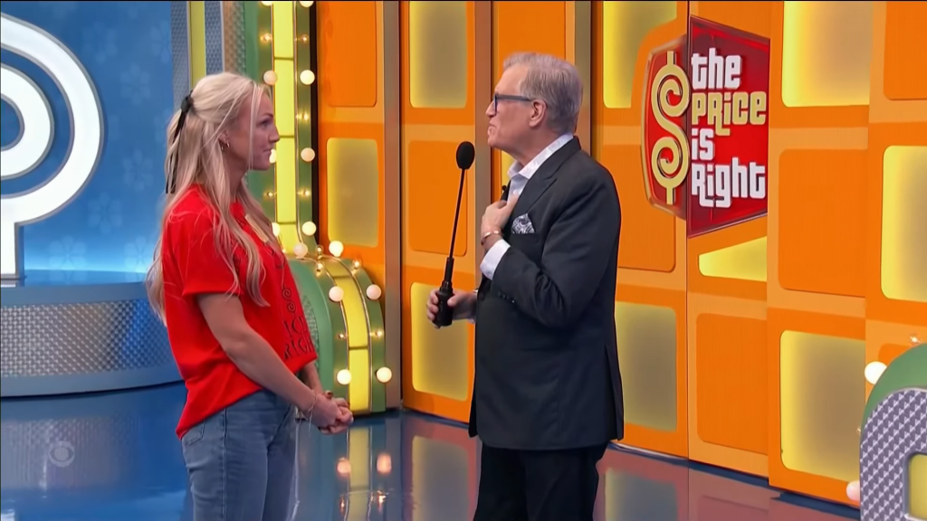 A contestant in a red shirt stands smiling in front of host Drew Carey on the set of 'The Price is Right', with colorful game show backdrops.