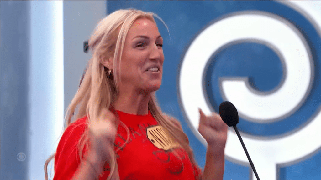 A smiling woman with long blonde hair, wearing a red shirt, excitedly raises her fists in celebration while standing in front of a blue backdrop with a large circular design.