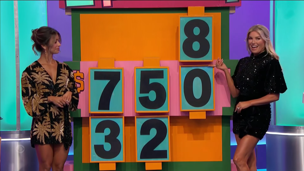 Two women standing beside a large colorful game board displaying the numbers 8, 7, 5, 0, 3, and 2, with one woman cheerfully pointing at the number 0.