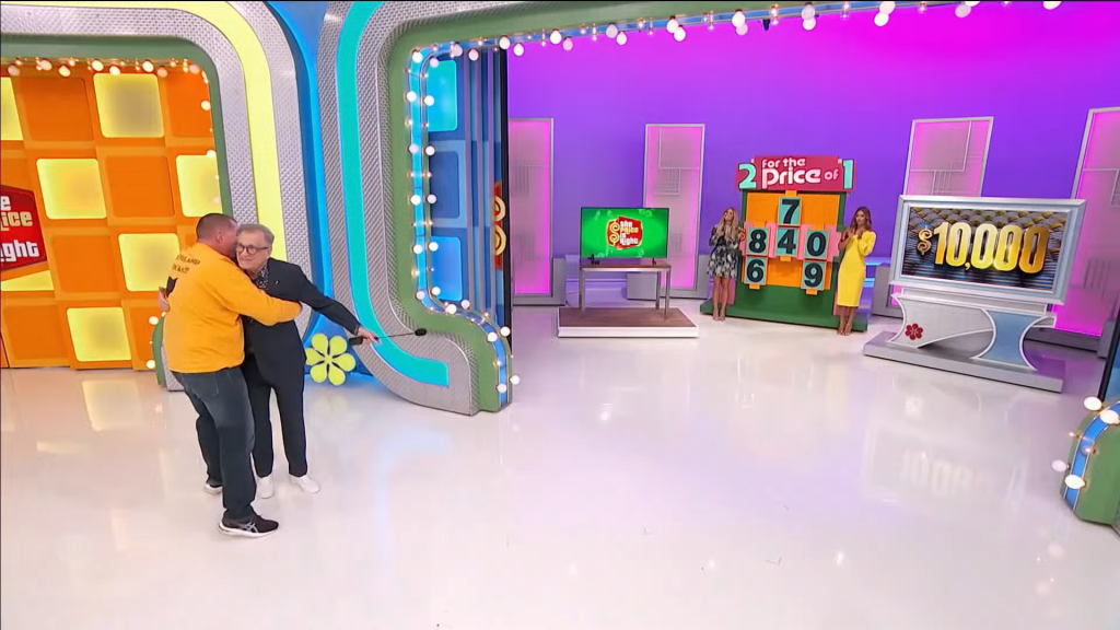 A contestant embraces Drew Carey on stage at The Price is Right, while behind them is a colorful game board displaying numbers and a prize total of ,000.