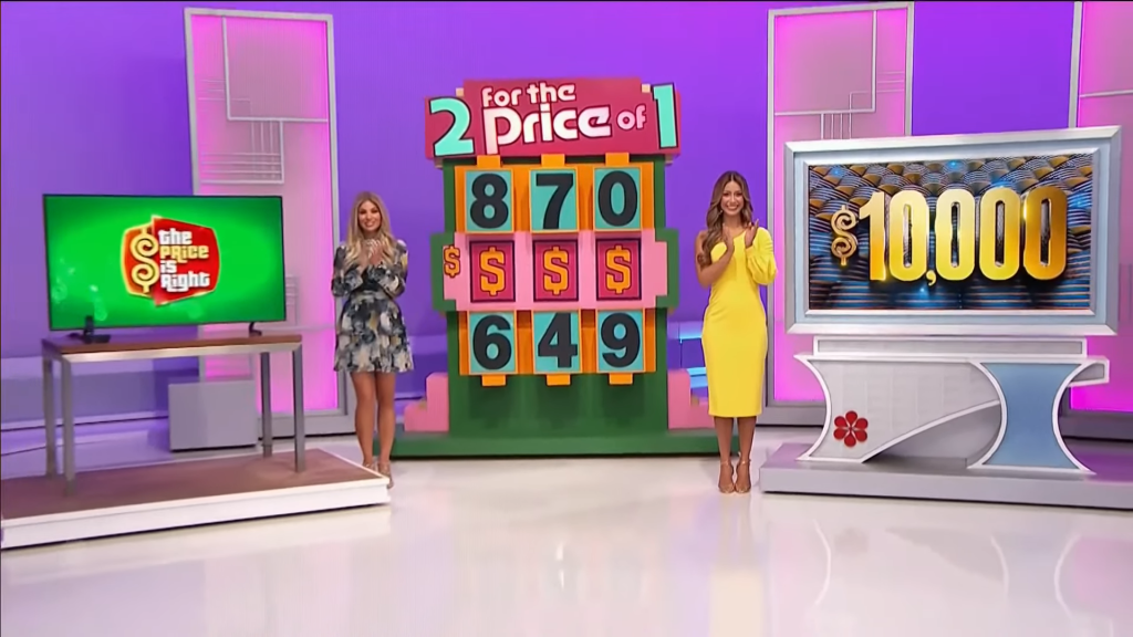 A game show set from 'The Price is Right' featuring two models, one in a floral dress and the other in a yellow dress, standing beside a colorful game board displaying the numbers 870 and 649, with a large ',000' sign in the background.
