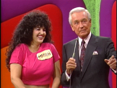 A woman with curly hair in a pink outfit bounces alongside a man in a suit holding a microphone, both appearing on the set of The Price is Right.