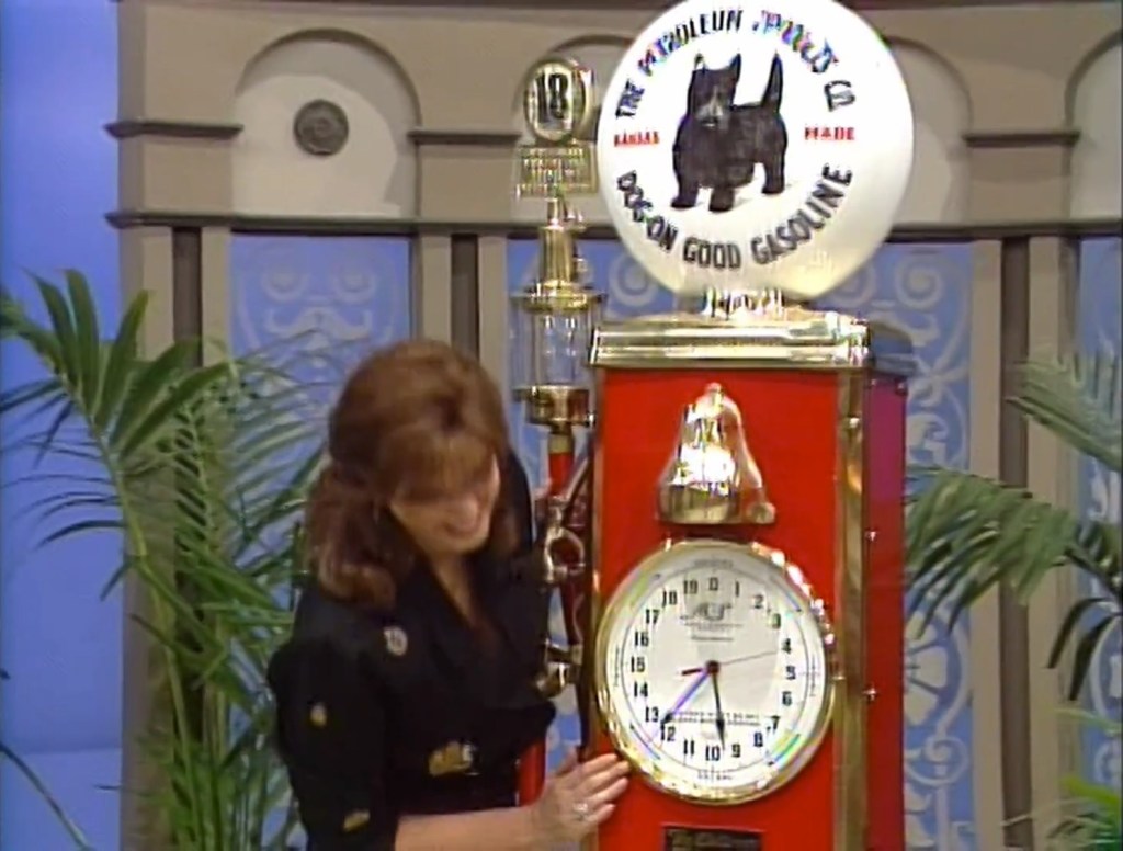 Holly Hallstrom interacts with a vintage red gas pump featuring a round glass globe that displays the image of a Scottish terrier and the text 'Doggone Good Gasoline.' The pump has a clock face and is set against a backdrop with decorative elements.